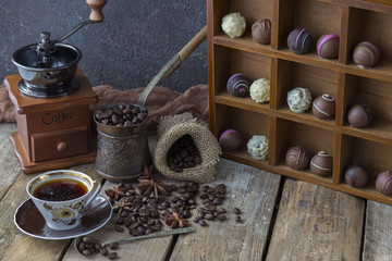 on a wooden table a coffee grinder, a Turk, a cup of coffee, a coffee grains and a cupboard with chocolates