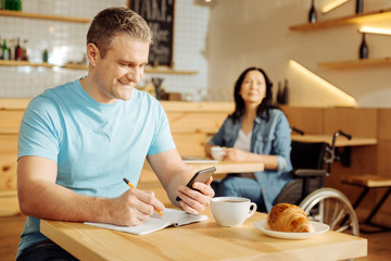 Distance working. Attractive joyful well-built blond man holding his phone and writing in his notebook while a woman sitting in a wheelchair in the background