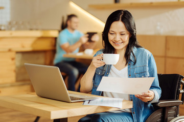 Reading. Beautiful alert dark-haired disabled woman sitting in a wheelchair and reading a document and drinking coffee