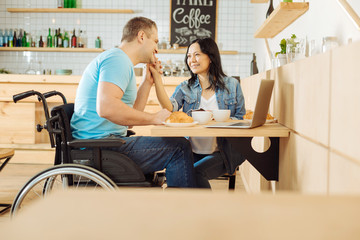 Leisure. Beautiful joyful dark-haired woman and a handsome smiling handicapped man sitting at the table in a cafe and listening to music and having coffee