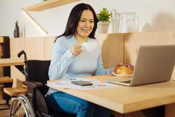Tea break. Beautiful happy dark-haired crippled woman sitting in a wheelchair and holding a cup of...