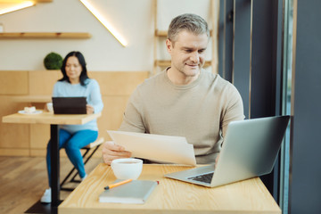 Fototapeta premium Working weekend. Attractive alert handicapped man sitting in a wheelchair and holding some sheets of paper and working on his laptop in a cafe and a woman sitting in the background