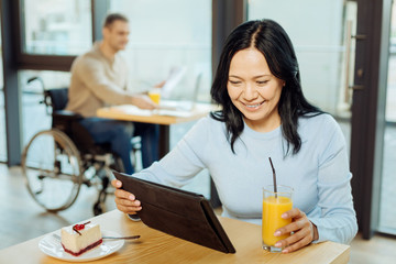 Piece of cake. Beautiful alert dark-haired woman smiling drinking juice and using her tablet while sitting in a cafe and a man in the wheelchair sitting in the background