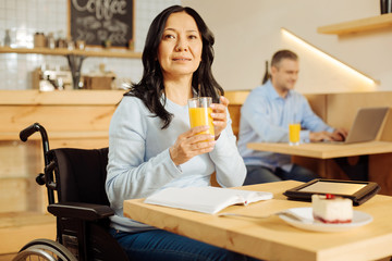 Healthcare. Pretty vigorous disabled woman sitting in a wheelchair and drinking juice and looking in the distance and a man sitting in the background