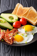 fried eggs with bacon, green onions, fresh avocado, toast and tomatoes close-up on a black plate. vertical