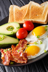 Homemade fried eggs with bacon, onion, avocado, toast and tomatoes close-up on a black plate. vertical