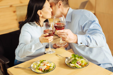 To us. Joyful disabled dark-haired woman and a content blond well-built man sitting in a cafe and kissing and drinking wine
