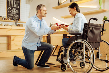 Be my wife. Attractive content well-built man smiling and proposing to his beloved dark-haired handicapped woman and holding a ring while having romantic dinner