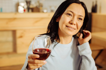 Nice day. Attractive happy dark-eyed woman smiling and looking in the distance while drinking red wine