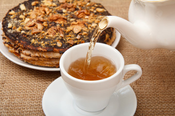 Tea is poured into a cup from porcelain teapot with homemade chocolate puff cake on the background