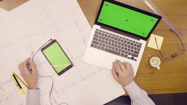 Top view. Man in headphones looking for something in tablet with green screen. A laptop with green screen laying on a table. Architect working at a desk, use laptop and tablet in the office