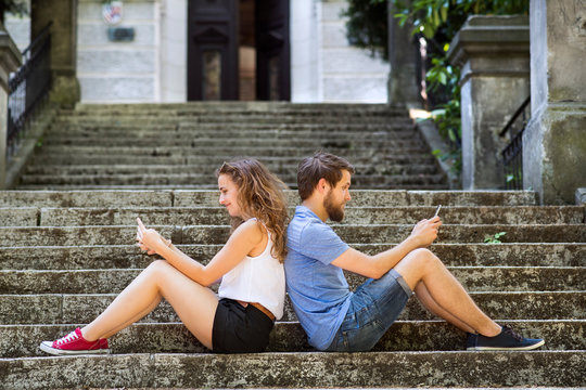 Young couple with smartphones sitting on stairs in town.