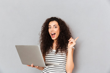Studio portrait of woman with curly hair being excited to find useful information in internet via silver computer gesturing eureka with index finger