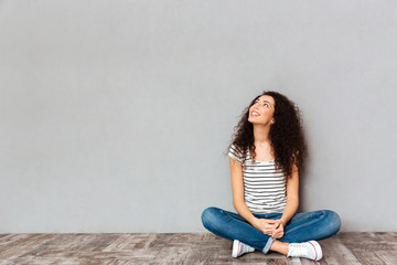 Young female in casual clothes sitting in lotus pose on the floor looking aside posing over grey wall copy space