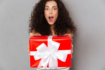 Fototapeta premium Brunette happy woman in defocus posing against grey wall holding red box gift-wrapped with white bow showing it on camera