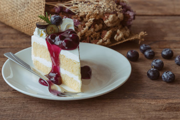 Blueberry cream cake on white plate on wood table in side view. Vanilla sponge cake decorated with dairy whipped cream and blueberry sauce so soft sweet and delicious. Homemade bakery concept.