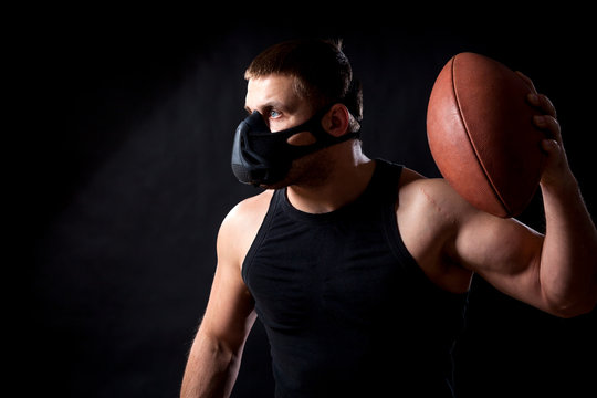 A dark-haired male athlete in a black training mask, a sports shirt holding a rugby ball and waving it on a black isolated background - Powered by Adobe