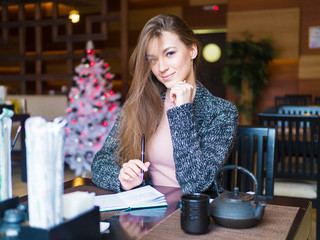 Beautiful successful young business woman enjoying creative job in cafe interior solving project in good mood, smiling entrepreneur checking notes in dairy making schedule of daily work holding pen