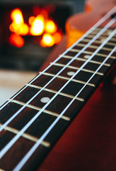 Fototapeta premium Ukulele small guitar close up stings, fireplace on the background. Musical concept, guitar fret board macro, fire in chimney, cosy romantic atmosphere.