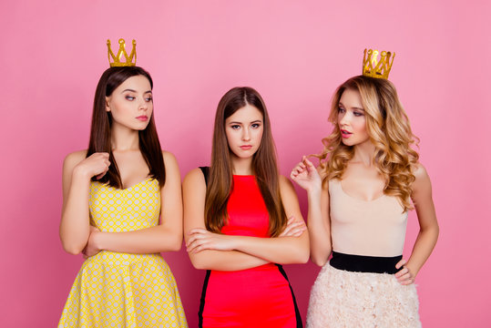 Two proud girls in gold crowns on head looking arrogantly to their unhappy disappointed friend without diadem with crossed hands, standing over pink background