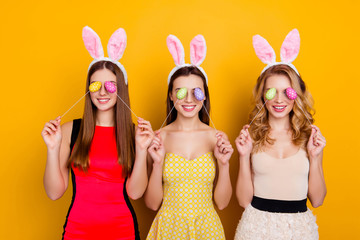 Happy Easter! Three pretty, trendy, comic, funny girls wearing bunny ears on heads in dresses closed eyes with easter eggs, standing over yellow background