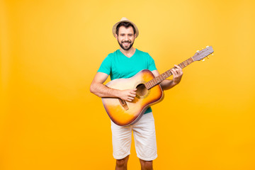 My guitar is my second soul! Portrait of smiling joyful funky glad positive guy with bristle, wearing tshirt and shorts, he is playing acoustic guitar, isolated on yellow background