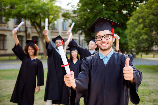 Happy caucasian male grad is showing good sign, blurred class mates are behind. He is in a black mortar board, with red tassel, in gown, with black spectacles, diploma in hand