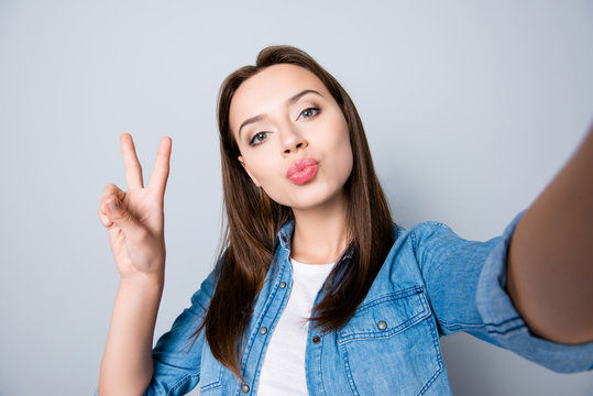 Self-portrait of confident brunette cute girl  blowing kiss to the camera and gesturing peace symbol, standing over grey background