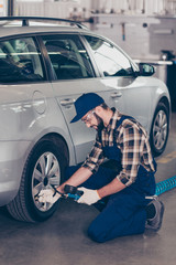 Bearded specialist, technician expert in protective glasses, blue overall, checkered shirt is analyzing tire physical pressure of silver car on hardware in workstation