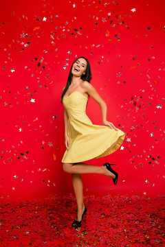 Vertical full-size full-length portrait of charming glamorous stylish fancy elegant shiny classy cheerful joyful attractive woman holding bottom on the dress dancing under confetti on red background