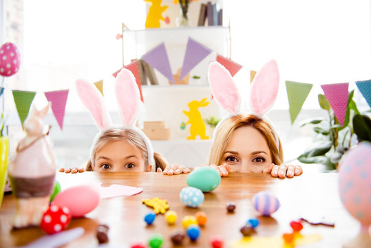 Two cute little sisters, friends wearing bunny ears, rabbit costumes, playing egg hunt on Easter, adorable child with mother celebrate Easter at home, looking out table
