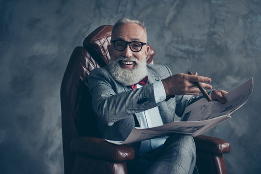 Attractive, rich, old, laughing boss with beaming smile in glasses, tux with red bow, have, hold newspaper, smoke cigarette, sitting in leather armchair in workplace over gray background, relax, fun