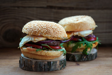 two hamburgers buns with sesame seeds, arugula, egg, cutlet lies on the stand of natural wood on a dark background