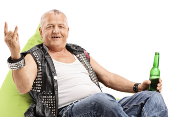 Old punker with a beer bottle seated on a beanbag making a rock hand gesture