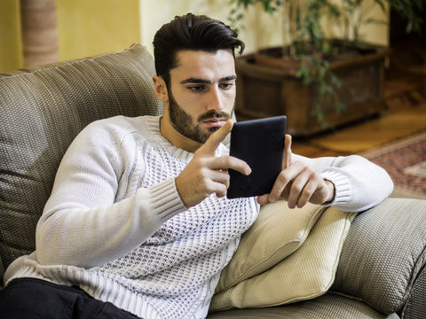 Handsome young man at home smiling, reading with ebook reader lying on a couch