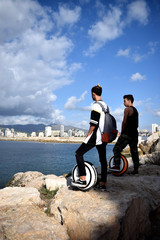 Two boys with mono wheels on the beautiful scenery of the Mediterranean Sea against the backdrop of the city of Calpe, the mountains and the blue sky with white clouds © Olga
