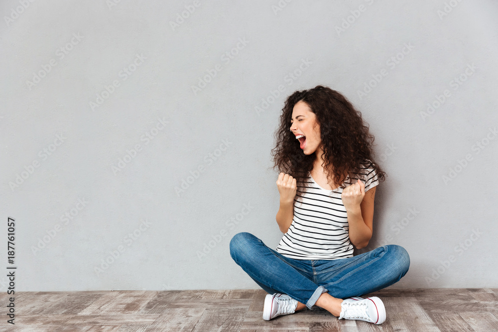 Poster young curly woman in casual clothes expressing delight while sitting in lotus pose on the floor clen