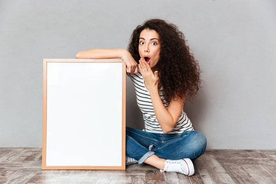 Funny portrait of adult girl in jeans sitting in lotus pose on the floor expressing embarrassment and surprise while holding picture in frame copy space