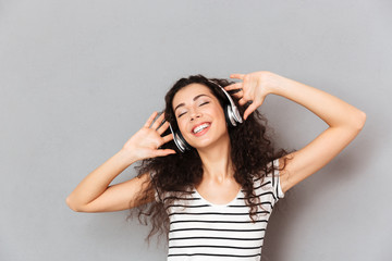 Picture of pleased young woman in striped t-shirt enjoying favourite songs listening melodies via headphones with closed eyes over grey background