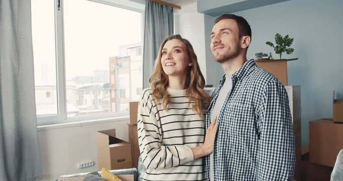Young beautiful couple in love standing in center of room and looking at new apartment. Guy kissing girlfriend on cheek. Smiling.