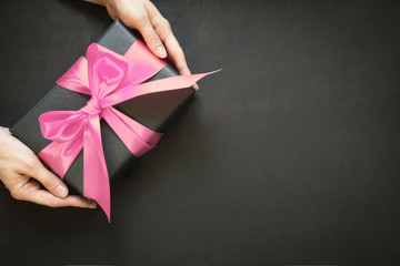 Gift box wrapped in black paper with pink satin ribbon in female hand on black surface. Copy space....