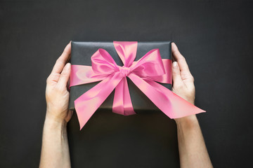 Gift box wrapped in black paper with pink satin ribbon in female hand on black surface. Copy space. View from above.