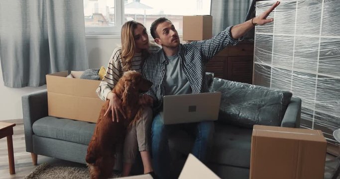 Young family sitting on couch. Guy holding laptop in hands. Pretty girl stroking dog. Happy couple talking about new purchases.
