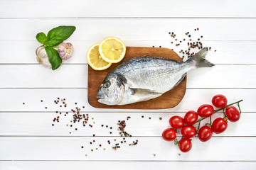 Fresh dorado fish on wooden cutting board with tomatoes, lemon, basil and garlic  on white wooden table. Top view, copy space.