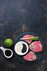 Fresh uncooked tuna fillet with condiments on a stone slate. Dark brown stone background, vertical shot with copyspace, flat-lay