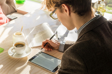 Doing home assignment project at cafe during lunch. Young modern male teenage student works with tablet pc and books on bright sunny day in cozy restaurant