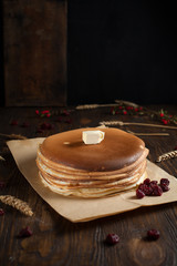 a pile of pancakes, berries, cookies, butter and a cutting board on parchment on a dark wooden table