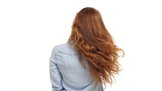 Back view of young ginger woman in denim shirt playing with her hair and looking at camera over white background