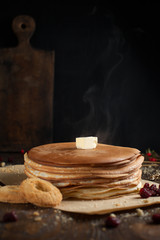 a pile of pancakes, berries, cookies, butter and a cutting board on parchment on a dark wooden table