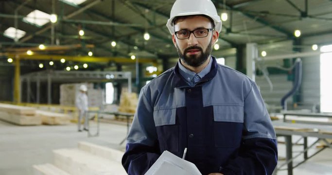 Portrait shot of factory worker ina hemlet and glasses standing with papers in the big furniture factory werehouse and posing into the camera. Indoors